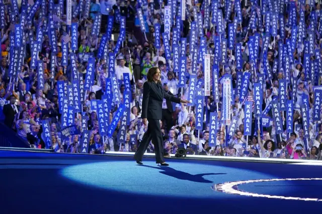 Kamala Harris sorrindo ao caminhar no palco; atrás, dezenas de democratas levantam faixa com seu nome 