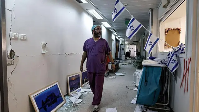Bir İsrailli doktor hasar alan hastanenin koridorunda yürüyor, üzerinde önlük var ve koridora İsrail bayrakları asılmış.