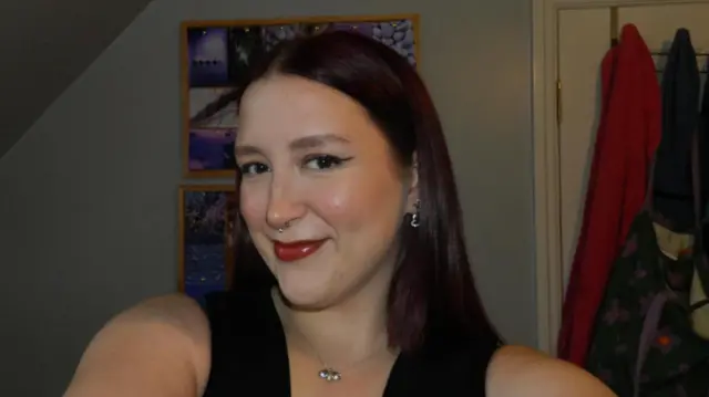 Selfie photograph of Tilly Short who smiles at the camera. She has dark brown hair, and wears red lipstick. She has a silver necklace on and a silver nose ring. 