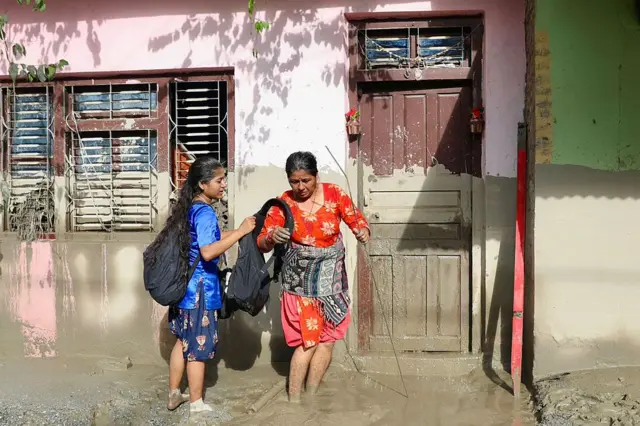 நீரின் அளவைக் காட்டும் சேற்று அடையாளங்களைக் கொண்ட வீடொன்றின் அருகில் பெண்கள் நடந்து செல்லும் காட்சி.