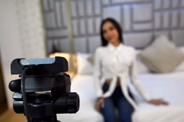 Una muchacha posa ante una cámara web vistiendo una camisa blanca y unos blue jeans. Ella está desenfocada, y está sentada sobre una cama.