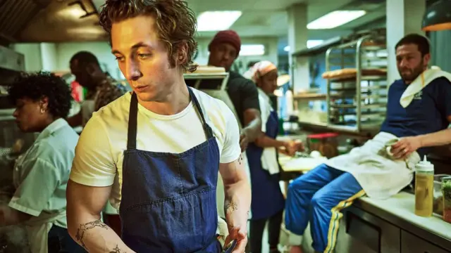 A foto mostra um homem jovem de pele e cabelos claros, com uma camiseta branca e um avental azul escuro em uma cozinha.