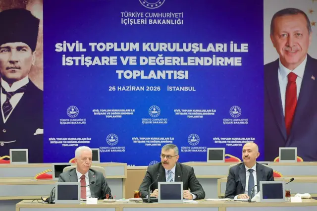 Üç takım elbiseli erkek yan yana oturuyor. Önlerinde ekranlar var. Arkada Erdoğan ve Atatürk resimleri. Panoda "İçişleri Bakanlığı Scil Toplum Kuruluşları ile İstişare Toplantısı" yazıyor. 