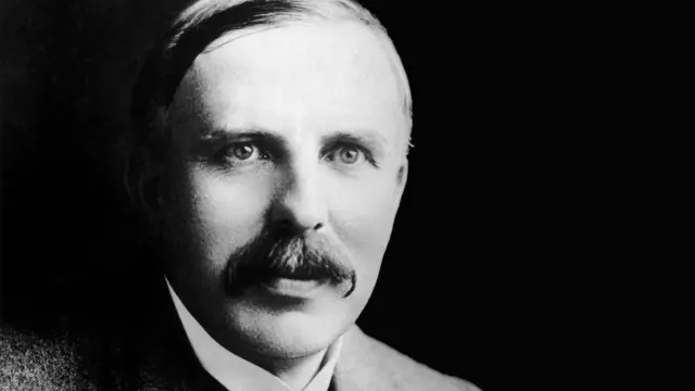 Ernest Rutherford, com um grande bigode, cabelo repartido para o lado e um colarinho branco rígido, olha além da câmera