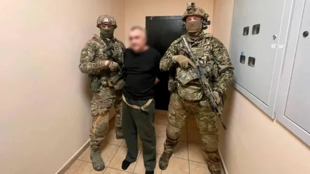 Двое вооруженных людей в военной форме с закрытыми маской лицами и задержанный в черной одежде, лицо которого заблерено