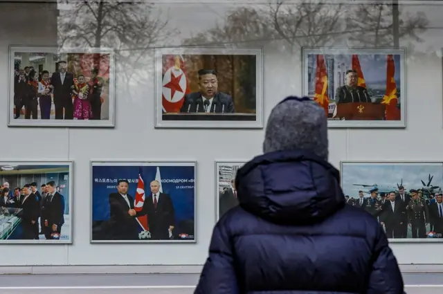 A man in a blue puffer jacket and a grey hat watches a series of photos of North Korea's leader, Kim Jong Un, through a window