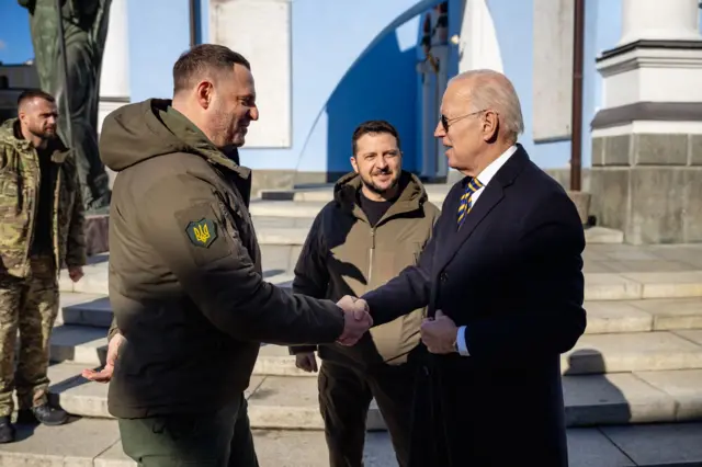 Єрмак у оливковій куртці тисне руки Джо Байдену у чорному пальто. За ними стоїть Зеленський і посміхається 