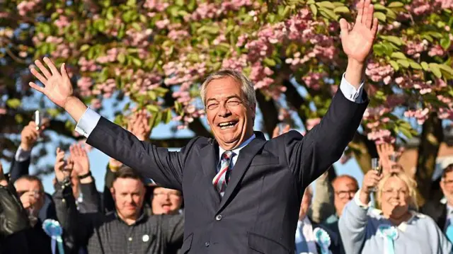 Найджелл Фарадж в костюме широко улыбается с поднятыми вверх руками, на его фоне люди поднимают бокалы возле цветущего дерева