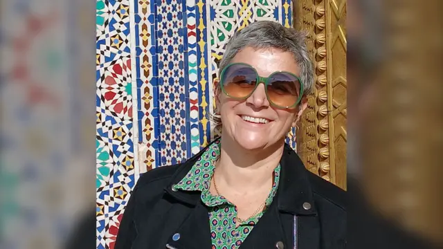 Raquel está de pie frente a una pared de mosaicos, sonriendo. Tiene el pelo corto y lleva unas gafas de sol grandes, redondas y verdes. También lleva una camisa verde estampada con una chaqueta negra encima.