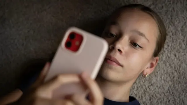 A teen wearing a navy shirt looks at her phone screen, her pink phone partially covering her face