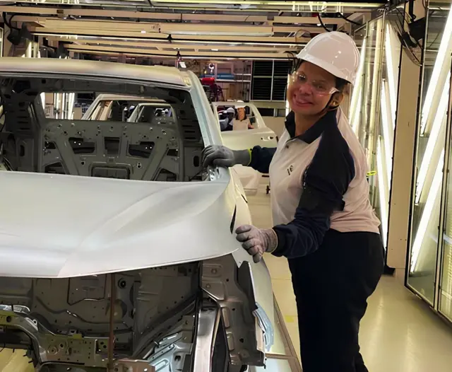 Gabriele de Oliveira Pereira usa capacete branco, uniforme de trabalho e luvas e sorri, enquanto apoia as mãos em um veículo ainda em processo de montagem na linha de produção da GWM em Iracemápolis, no interior de São Paulo