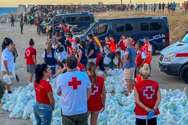 Personal de la Cruz Roja está parado frente a insumos en bolsas que serán repartidos entre muchas otras personas que esperan en fila en una playa de Ceuta. Se ven también furgones de policía y personal de ayuda.