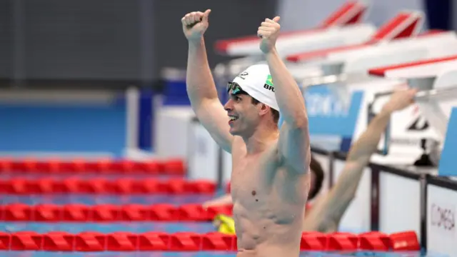 A foto mostra um nadadorquantos saques posso fazer na greenbetsuma piscina, usando uma toucaquantos saques posso fazer na greenbetsnatação branca, com os braços para cima comemorando.