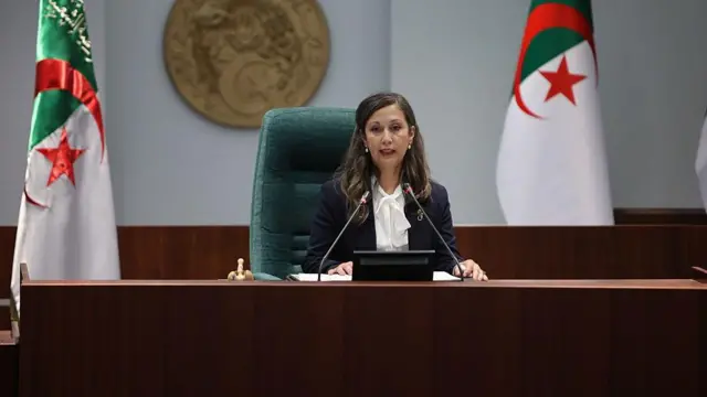 خليدة بوفدش على كرسي رائسة مجلس الشعب تلقي كلمة وبجانلها علم الجزائر