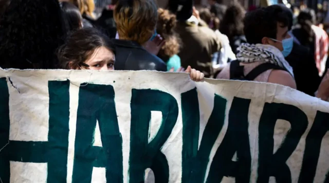 Harvard Üniversitesi'ndeki Filistin yanlısı protestolardan birinde bir kız, yeşil renkli Harvard yazısının arkasından endişeli gözlerle bakıyor. Sadece gözleri var. Arkada Filistin yanlısı başka gençler de duruyor.