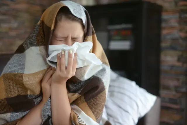 একজন নারী কম্বল গায়ে পেঁচিয়ে ও মুখে টিসু ধরে হাঁচি দিচ্ছেন