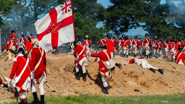 Homens com uniformes vermelhos e brancos sobem uma duna durante uma encenação de batalha