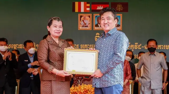 A man and a woman both hold a framed certificate and smile at the camera, while a row of people wearing face masks stand behind them