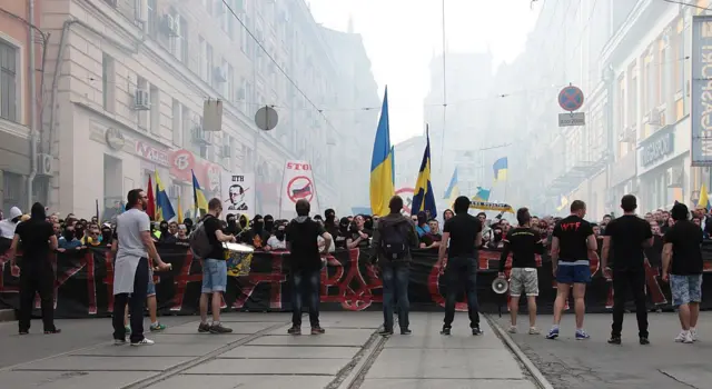 Великий натовп людей на міській вулиці з українськими прапорами та плакатами. Попереду – ряд учасників із великим чорним банером з червоним тризубом. На задньому плані – густий натовп із прапорами та гаслами, у повітрі легкий дим.