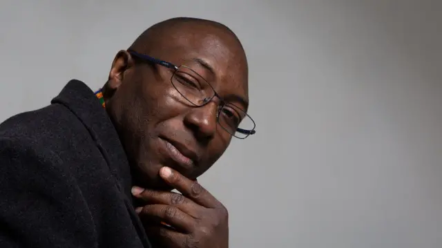 Dominic Nelson-Ashley looking into the camera with his hand resting on his chin. He is wearing glasses and a black coat.