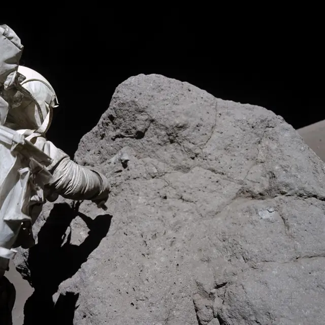 O astronauta Harrison Schmitt, da Apollo 17, coleta amostras de rochas da Lua. A rocha é um pouco mais alta que ele e tem cerca de três vezes sua largura. Ele está à esquerda da foto, de costas para a câmera, vestindo um traje espacial branco da Nasa, e tenta coletar a amostra usando seu braço direito. Seu corpo cria uma sombra sobre a grande rocha cinza escura.

