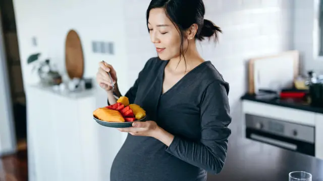 Молода вагітна азіатка їсть свіжі та різнокольорові органічні фрукти вдома
