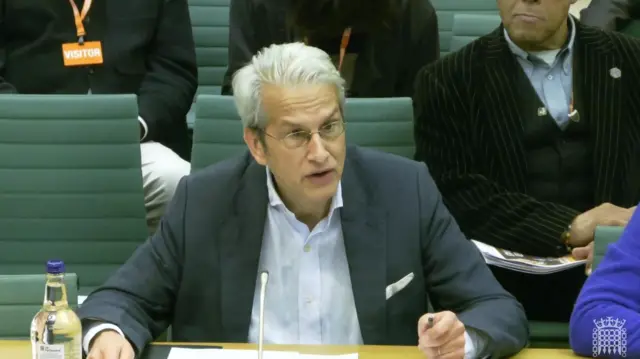 白髪をオールバックにして眼鏡をかけ、灰色の上着に白いシャツを着た男性が、机に向かって座り、マイクを前に話している。後ろには緑色の椅子が並ぶ