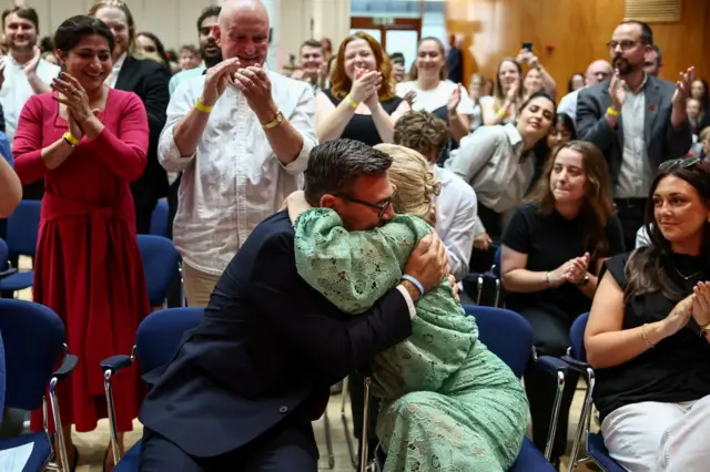 ホールに並べられたパイプ椅子に隣同士に座るバーナム氏とヴァン・ヒール氏が抱き合っている。ヴァン・ヒール氏は緑色のワンピースを着ている。後方の席の人々が、その様子を立って拍手をしながら称えている