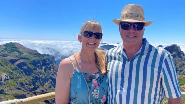 Lee con una camisa azul y blanca a rayas, un sombrero canotier de paja y gafas de sol, de pie junto a su esposa, que lleva un vestido de verano floreado de color turquesa y gafas de sol, en un balcón, en lo alto con un impresionante telón de fondo de picos de montañas, nubes bajas y cielos azules detrás de ellos.