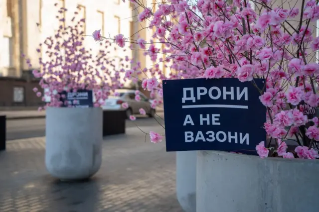 Вуличні бетонні вазони зі штучними рожевими квітами; на одному з них табличка з написом "ДРОНИ, А НЕ ВАЗОНИ". У тлі – міська вулиця.