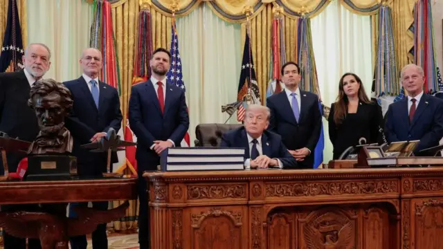 トランプ氏が机の前のいすに座り、その背後でスーツ姿の人々が両側に並んでいる。全員、同じ方向を向いている