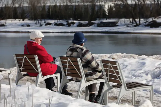 Две женщины сидят на скамейках на заснеженном берегу реки