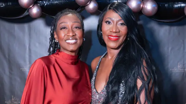 Beverley Knight, con el pelo negro trenzado, sonríe a la cámara. Lleva un vestido rojo de seda. Pauline Reid, a su lado, tiene el pelo largo y negro, un vestido plateado con piedras preciosas y pintalabios rojo.