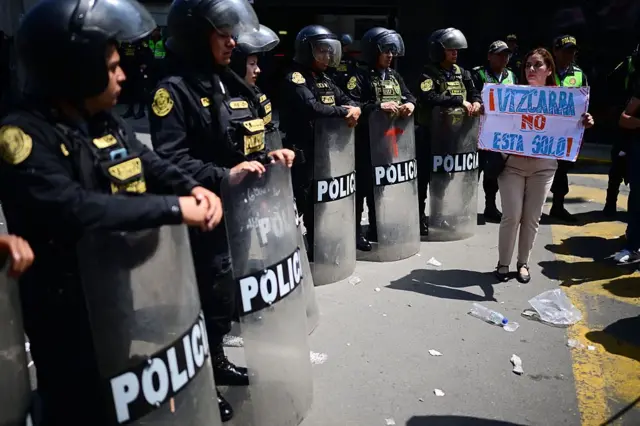 Una manifestante sostiene un cartel en el que dice "Vizcarra no está solo" frente a una línea de policías antimotines, quienes sostienen escudos plásticos. 