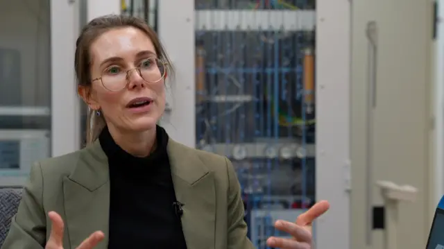 Una mujer con chaqueta caqui, jersey negro de cuello alto y gafas redondas habla a una cámara descentrada, gesticulando con las manos. Está sentada en un laboratorio con varios equipos ocultos tras ella. Lleva el pelo oscuro recogido.