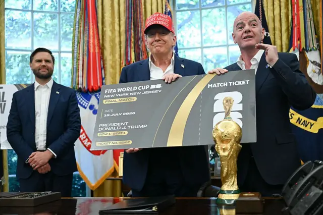 Fotoğraf, Amerika Birleşik Devletleri'nin Washington şehrindeki Oval Ofis'te çekilmiş. ABD Başkanı Donald Trump ve FIFA Başkanı Gianni Infantino, 2026 Dünya Kupası Finali biletini tutarken görülüyor. Her ikisi de takım elbise giymiş, Trump ayrıca önünde "Trump her konuda haklıydı" yazısı işlenmiş kırmızı bir MAGA şapkası takıyor. Arka planda JD Vance görülebiliyor. Tüm adamlar bir masanın arkasında duruyor, ön planda ise Dünya Kupası kupası yer alıyor.