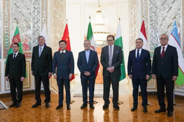 شرکت کنندگان نشست تهران مردانی با دریشی مقابل پرچمهای روسیه، چین، پاکستان، تاجیکستان، ازبیکستان، ایران و ترکمنستان ایستادهاند