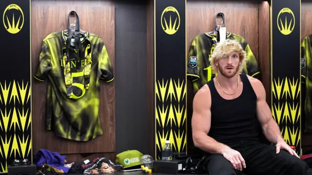 Logan Paul, homem musculoso de cabelo loiro platinado, veste uma regata preta enquanto está sentado em um vestiário