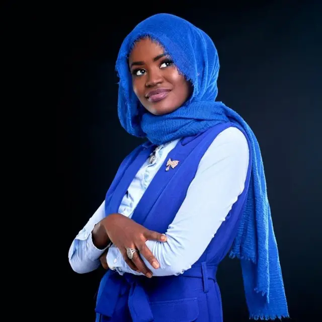 Portrait de la coach Mariama Ndiaye. Elle porte un voile bleu, et une combinaison assortie avec une chemise bleue ciel, sur un fond noir.