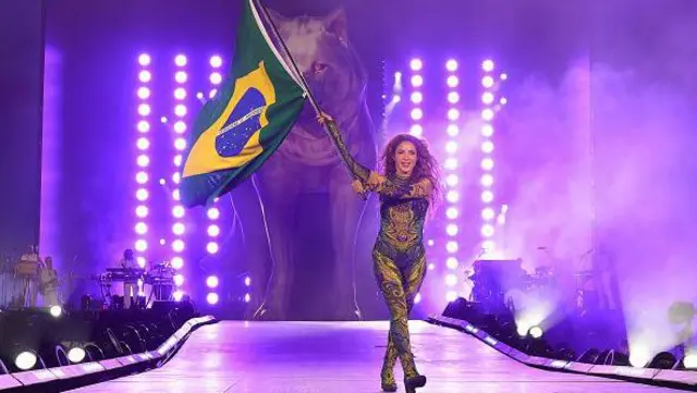Shakira no palco em Copacabana, segurando uma bandeira do Brasil e com uma estátua de um lobo ao fundo