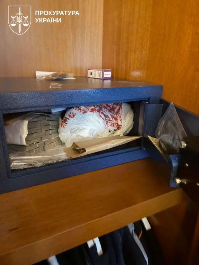Відчинений невеликий сейф, вбудований у шафу з дерев’яними стінками.
Усередині лежать пачки готівки, паперові конверти та пакет, поряд зверху видно коробку з-під сірників і дрібні предмети.
