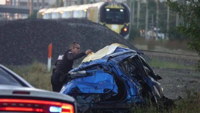 Un agente de policia de Aventura examina los restos de un auto destrozado tras una colisión con un tren Brightline. En segundo plano, un tren de la línea. 