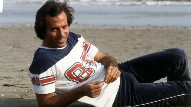 Хуліо Іглесіас, значно молодший, позує з посмішкою, лежачи на березі моря