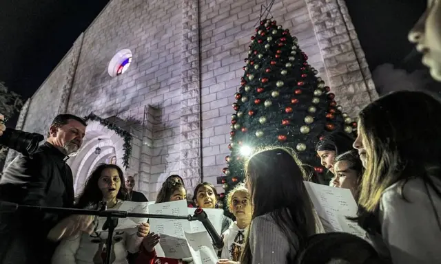 Children and adults singing in front of a Christmas tree and church