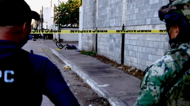 Солдат та ще один чоловік у формі стоять за стрічкою поруч із місцем вбивства, далі на тротуарі, поруч зі своїм велосипедом лежить мертвий хлопець