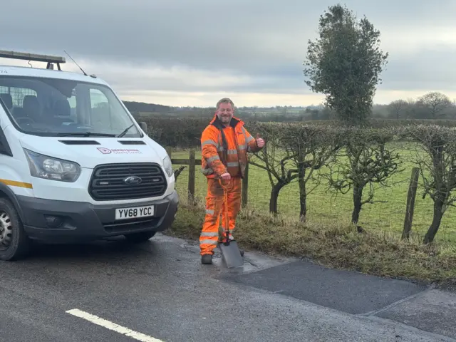 Un miembro de un equipo de reparación de carreteras, con pantalones y chaqueta naranja brillante con franjas reflectantes, está de pie, con el pulgar izquierdo levantado y una pala en la mano derecha. Está delante de una furgoneta blanca del Consejo del Condado de Derbyshire, junto a un tramo de carretera recién reparado.