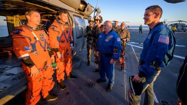 オレンジ色の宇宙服を着た2人が笑顔で、輸送艦の甲板でヘリコプターを背に立ち、夕日を浴びている。青いジャケットを着たNASA長官や他の関係者が笑顔で二人の前にいる