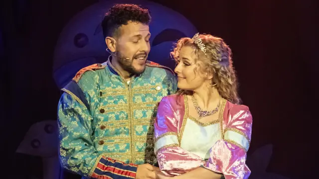 Alicia está a la derecha con un vestido de terciopelo rosa y blanco. Tiene el pelo rubio y rizado y lleva una tiara. Mira de reojo a un hombre que viste una chaqueta verde azulado con detalles dorados. Tiene el pelo castaño.