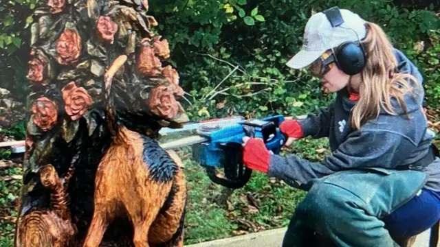 Kimberly Hone construye el banco con una motosierra. Lleva ropa protectora, gorra y gafas de seguridad. Está agachada, trabajando en el diseño de un perro.