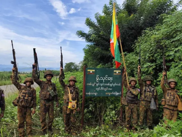 မြန်မာအလံထောင်ပြီ ရွာဆိုင်းဘုတ်နားမှာ မတ်တပ်ရပ်နေကြတဲ့ စစ်သား၆ဦး